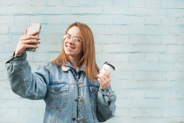Hermosa chica feliz se encuentra en el fondo de una pared azul con una taza de café en las manos y toma una selfie en un teléfono inteligente. Hipster chica hace una selfie, lleva una chaqueta jeans y gafas . — Foto de Stock