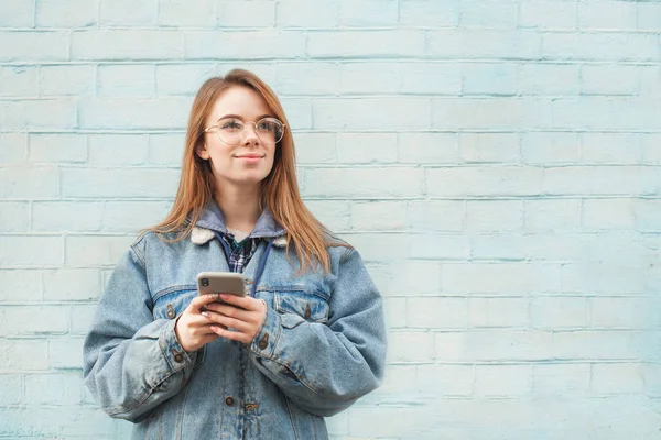 Χαμογελαστή κορίτσι των χίπλερς σε ένα ντένιμ μπουφάν και γυαλιά στέκεται στο φόντο ενός γαλάζιου τοίχου με ένα smartphone στα χέρια του και κοιτάζοντας πλάγια σε ένα κενό μέρος. Χώρος αντιγραφής — Φωτογραφία Αρχείου