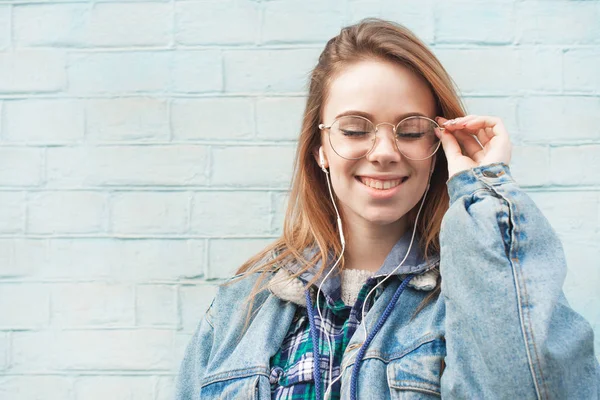 Close up retrato de uma menina atraente em uma jaqueta de ganga fica no fundo de uma parede azul nos fones de ouvido, ouve música com os olhos fechados e tem prazer. Espaço de cópia — Fotografia de Stock