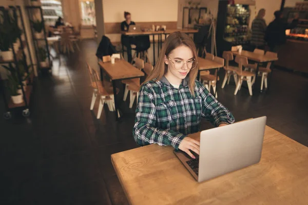 Gyönyörű fiatal lány dolgozik egy laptop egy hangulatos kávézóban, nézte a képernyőt, és mosolyogva. Egy vonzó női szabadúszó egy laptop ül egy kávézóban. Tanuló laptoppal tanul. — Stock Fotó