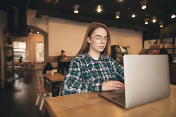 Menina atraente em óculos, vestindo uma camisa usa um laptop em um café acolhedor, está focado em olhar para a tela. Estudante aprende com um laptop no café . — Fotografia de Stock