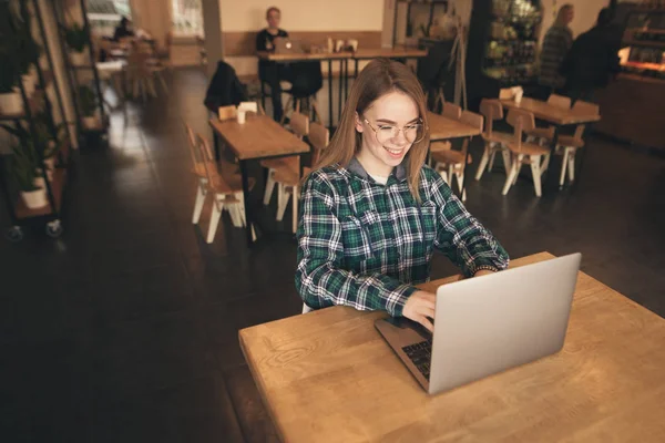 Retrato de un freelancer feliz se sienta en un acogedor café con un ordenador portátil, marca el texto en el teclado y sonríe. La bloguera positiva trabaja en un café con un portátil. Estudiante estudiando en un café . —  Fotos de Stock
