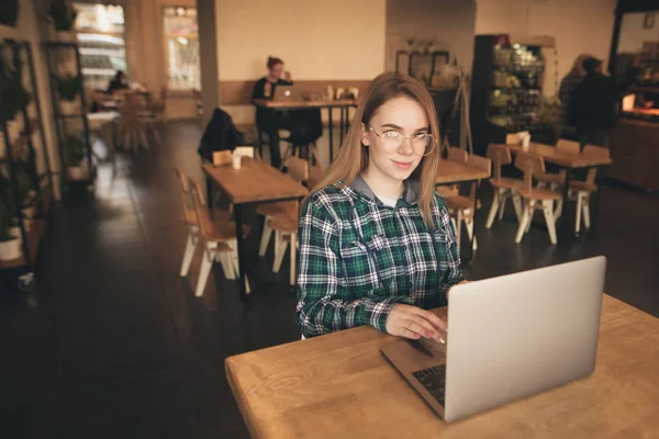 Mooi meisje gekleed in een shirt zit met een laptop in een gezellige koffieshop, werkt, kijkt naar de camera en glimlacht. Aantrekkelijk meisje student maakt gebruik van een laptop in een cafe. — Stockfoto