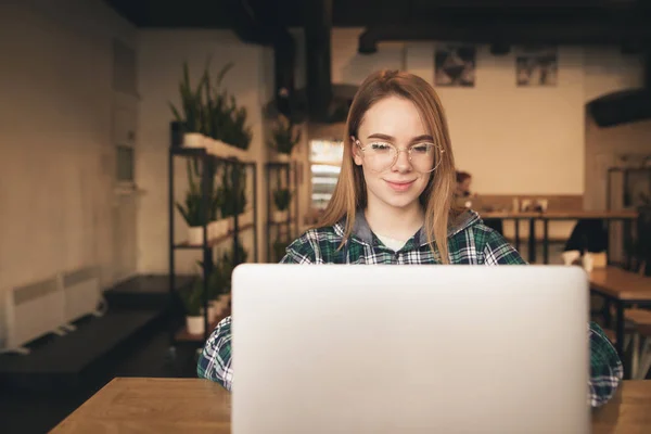 Muchacha atractiva trabaja en un ordenador portátil en un acogedor café, mira a la pantalla y sonríe. Hermosa chica blogger sentada con un portátil en la cafetería y sonriendo. Estudiante estudiando en un café . — Foto de Stock