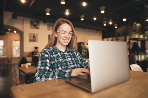 Retrato de un freelancer feliz se sienta en un acogedor café con un ordenador portátil, marca el texto en el teclado y sonríe. La bloguera positiva trabaja en un café con un portátil. Estudiante estudiando en un café . — Foto de Stock