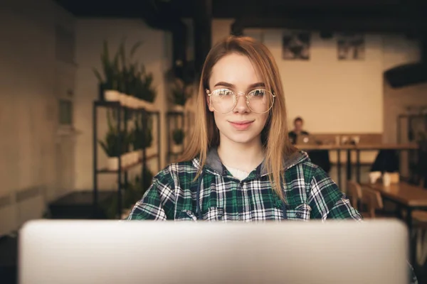 Retrato de uma menina atraente e positiva em um café acolhedor usa um laptop, olha para a câmera e sorri. Menina alegre em uma camisa e óculos em um café . — Fotografia de Stock