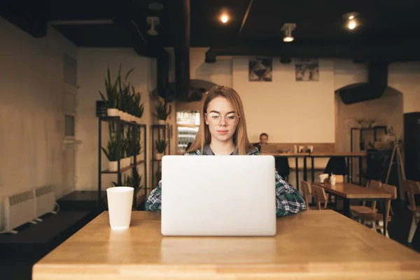 Menina freelance bonita trabalha em um café acolhedor em um laptop. Estudante menina em uma camisa estudando em um café, usando a Internet em um laptop. Trabalho na Internet . — Fotografia de Stock