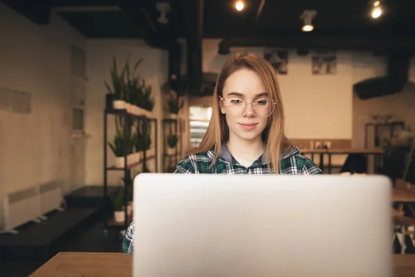 Muchacha atractiva con gafas y una camisa utiliza un ordenador portátil en un acogedor café, se centra en la pantalla y sonríe. Adolescente chica con portátil sentado en un café . — Foto de Stock