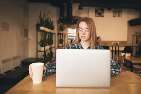 Hermosa chica trabaja en una cafetería en un ordenador portátil, toma una taza de café con su mano. Muchacha estudiante atractiva aprende en un acogedor café, utiliza Internet en un ordenador portátil . — Foto de Stock