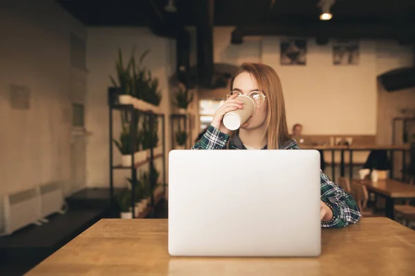 Lány egy alkalmi ruha ül egy kávézóban egy laptop az asztalra, és kávéfogyasztás. Fiatal nő dolgozik egy kávézóban egy laptop és italok teát a pohárból. — Stock Fotó