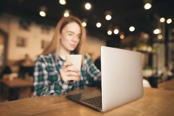 Háttér. Fiatal lány dolgozik egy laptop egy kávézóban, és tart egy pohár kávét a kezében. A nő egy laptopot használ a kávézóban. Fókuszban a laptop. — Stock Fotó