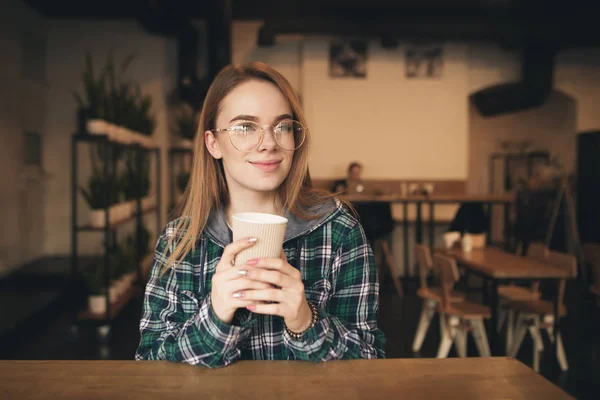 Счастливая студентка в очках и рубашке, сидит в кафе, держа чашку кофе в руках, смотрит в камеру и улыбается. Студентка отдыхает в кафе с чашкой кофе . — стоковое фото