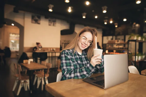 Retrato de uma menina sorridente em roupas casuais e óculos sentados em um café acolhedor com um laptop, posando na câmera. Menina feliz usa um laptop no café e olha para a câmera — Fotografia de Stock