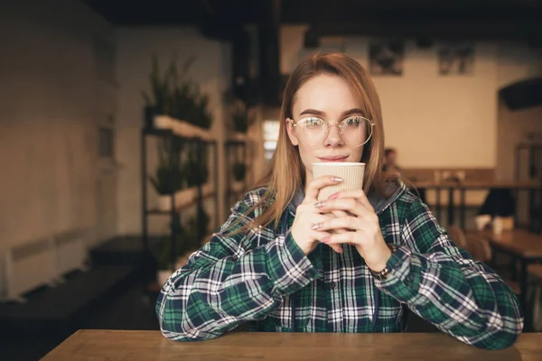 Chica estudiante feliz con gafas y una camisa, sentado en un café con una taza de café en las manos, mirando a la cámara y sonriendo. Estudiante descansando en un café con una taza de café . — Foto de Stock