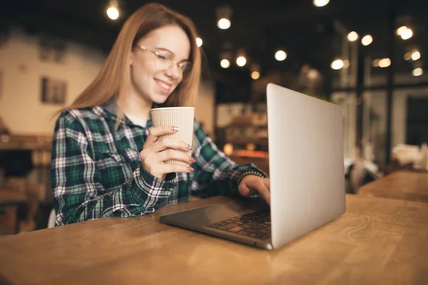 Menina feliz com um copo de café em suas mãos usa um laptop em um café, olha para a tela e sorri. Concentre-se em um copo de café na mão . — Fotografia de Stock