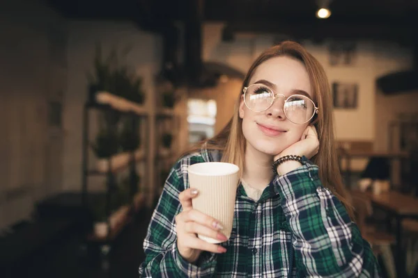 Fille attrayante rêveuse avec une tasse de café dans ses mains, portant des lunettes et une chemise, assis dans un café confortable, levant les yeux et rêves. Portrait d'un étudiant rêveur dans un restaurant . — Photo