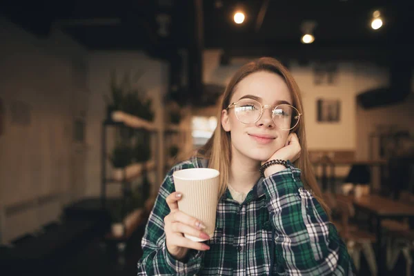 Retrato cercano de una hermosa chica sosteniendo una taza de café en su mano, mirando a la cámara y sonriendo. Estudiante positiva descansando en un café, tomando café en una cafetería . — Foto de Stock