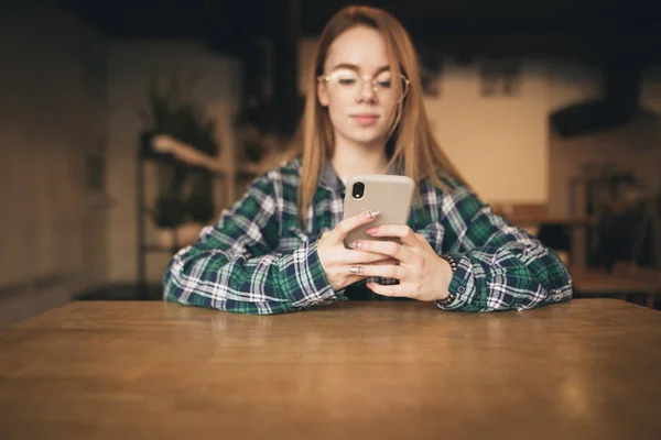 Smartphone en manos de una atractiva chica vestida con ropa casual, sentada en un acogedor café. Chica joven utiliza un teléfono inteligente, sostiene un teléfono en la mano y navega por Internet . — Foto de Stock
