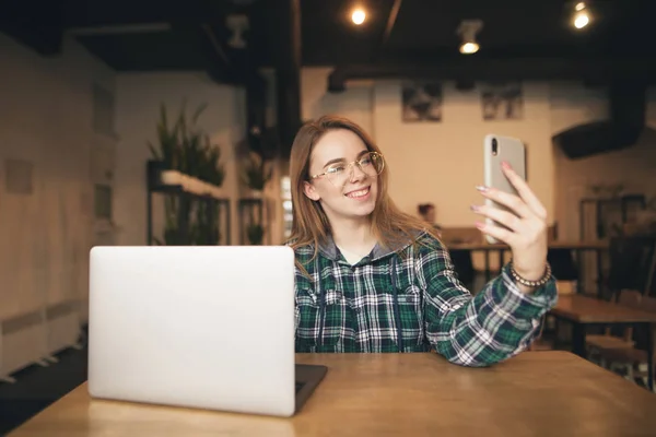 Vidám lány a szemüveg és egy alkalmi ruha ül egy kávézóban egy laptop, teszi selfie egy okostelefon, mosolygós és pózol. Lány szabadúszóként veszi selfie. Diák laptoppal és okostelefonnal — Stock Fotó