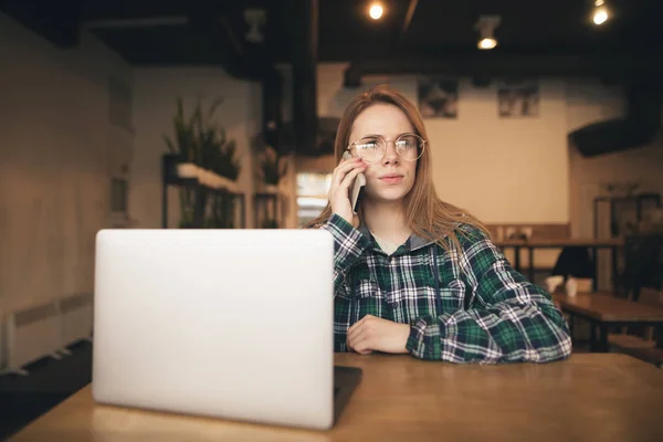 Menina atraente grave usa um laptop em um café acolhedor e fala por telefone, use roupas casuais. Jovem de negócios trabalhando em um café em um laptop, chamando o telefone . — Fotografia de Stock