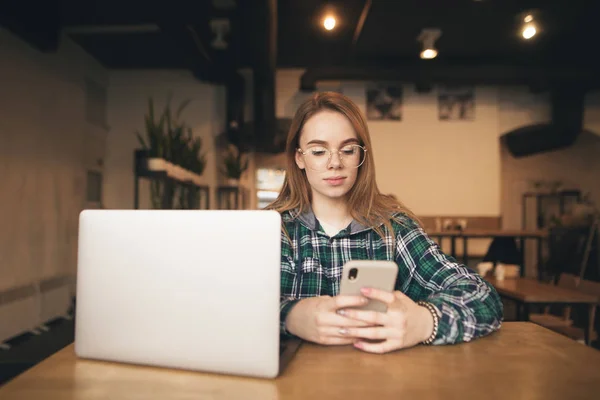 Retrato de uma menina atraente senta-se em um café acolhedor com um laptop e usa um smartphone, vestindo óculos e roupas casuais. Menina freelance focado trabalha em um café em um laptop e smartphone . — Fotografia de Stock