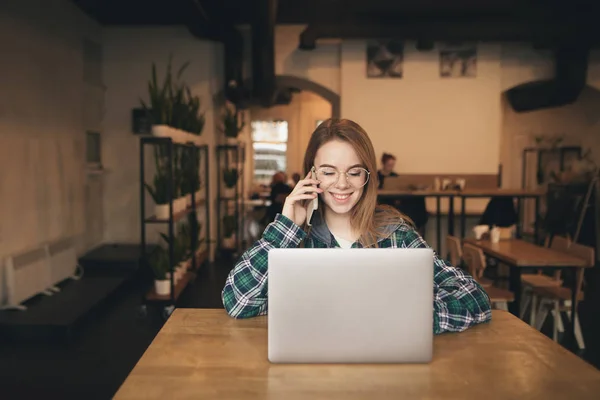 Smiling girl in casual clothes speaks by telephone and uses the internet on a laptop in a cozy cafe, look at the screen and smiles. Gadis ceria bekerja di kafe . — Stok Foto