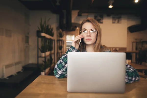 Schöne Mädchen telefoniert und arbeitet an einem Laptop in einem Café, trägt eine Brille und schaut weg. Student telefoniert mit Freunden und sucht Informationen im Internet, studiert — Stockfoto