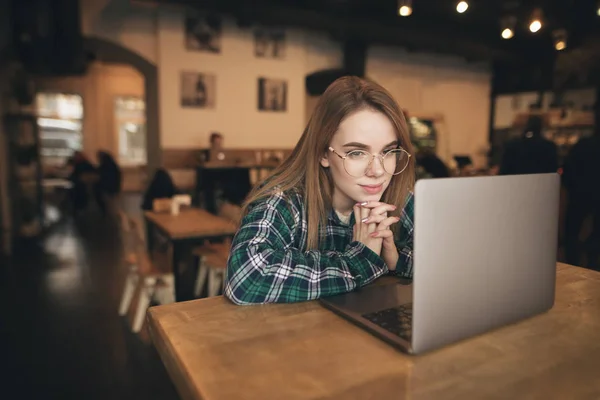 Rahat giysiler ve gözlük bir laptop ile rahat bir kafede oturan bir gülümseyen kız portre, kamera poz. Mutlu kız kahve dükkanında bir dizüstü bilgisayar kullanır ve kamera bakıyor