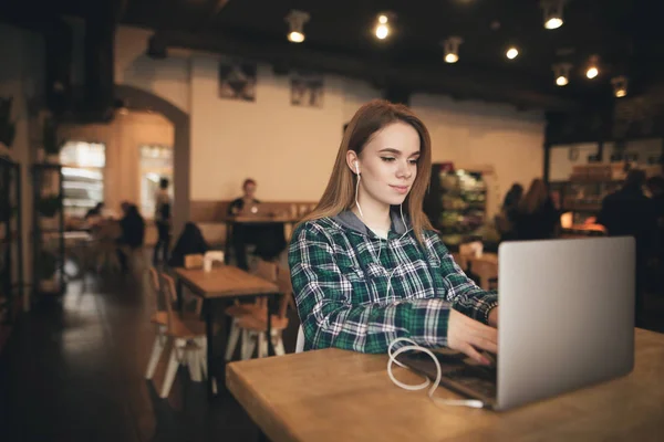 Menina bonito trabalha em um laptop em um café acolhedor e ouve música em fones de ouvido, usar roupas casuais, ela está focada em olhar para a tela. Freelancer trabalha em um café em um laptop . — Fotografia de Stock