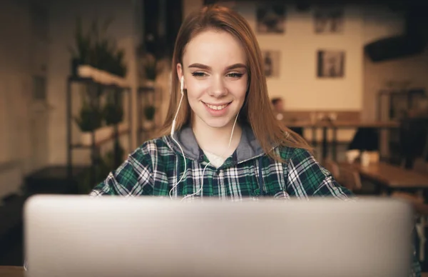 Menina sorridente em roupas casuais e fones de ouvido funciona em um laptop em um café, olha para a câmera e sorri. Conceito Freelance. Retrato de um estudante feliz em um café . — Fotografia de Stock