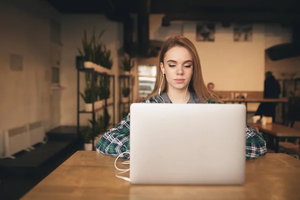 Estudante bonita goza de um laptop em um aconchegante café e está estudando. Retrato de uma menina atraente com um laptop no café, ouve música nos fones de ouvido e olha para a tela . — Fotografia de Stock