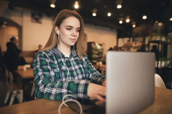 Muchacha atractiva estudiante se sienta en un café con un ordenador portátil, escucha música en los auriculares y funciona. Señora con un ordenador portátil en una acogedora cafetería . — Foto de Stock