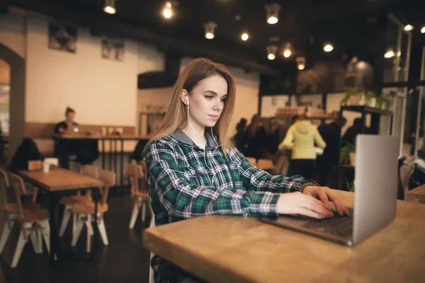 Retrato de una chica elegante usando una camisa, usando un portátil en la cafetería, mirando a la pantalla y escuchando el sonido en los auriculares. Hermosa chica trabaja en un ordenador portátil en un café . — Foto de Stock