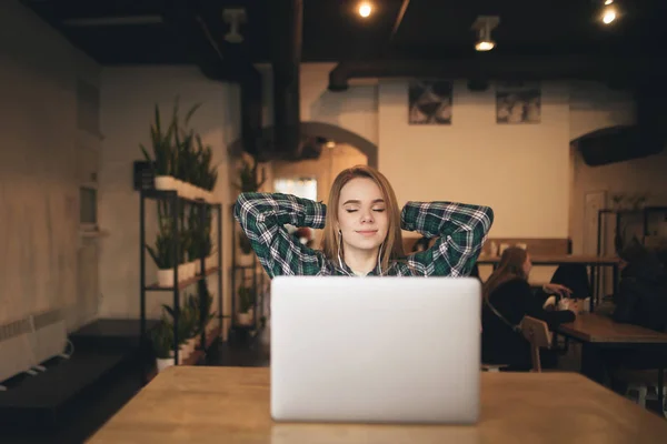 Menina atraente está tendo um descanso em um café com um laptop, ouve música nos fones de ouvido e relaxa com os olhos fechados. Menina estudante descansando em uma pausa em um café. Intervalo de freelance no trabalho . — Fotografia de Stock