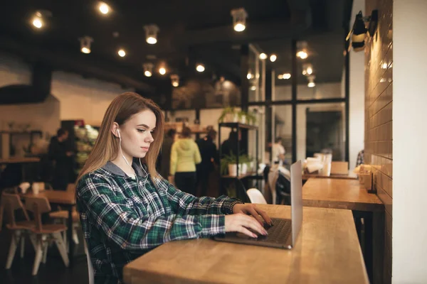 Schöne Studentin genießt einen Laptop in einem gemütlichen Café und studiert. Porträt eines attraktiven Mädchens mit Laptop im Café, hört Musik im Kopfhörer und blickt auf den Bildschirm. — Stockfoto