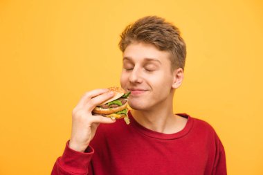 Gözleri ile memnun adam kapalı ve sarı bir arka planda elinde bir burger ile. Genç adam iştah açıcı bir burger kokur ve fast food yemek için memnun.