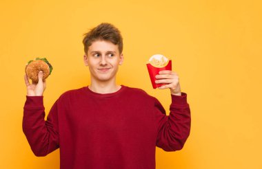 Rahat giysiler giyen komik adam patates kızartması ve elinde bir burger ile sarı bir arka planda duruyor. Aç genç adam, ellerinde, izole hızlı yiyecek tutar. Boşaltmak