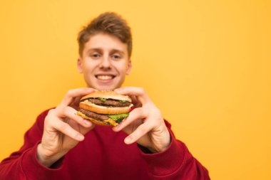 Elinde bir burger ile mutlu genç çocuk sarı bir arka plan üzerinde duruyor, Burger yakın çekim. Gülümseyen öğrenci elinde lezzetli bir burger tutar, sağ reklam için boş alan.
