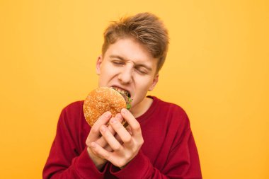 Gözleri sarı bir arka planda kapalı çok aç genç ısırma Burger yakın çekim fotoğrafı. Aç öğrenci hızlı yemek yiyor, portre yakın. Boşaltmak