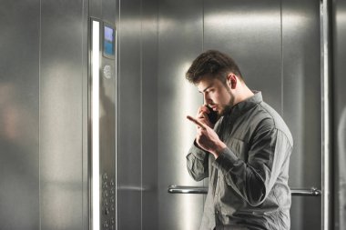 Adam asansörde gri gömlek giyen bir şeyi açıklıyor. Genç adam çalışan onun patron ile onu akıllı telefonda barışçı konuşuyor.