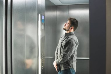 Sakallı adam asansörde duruyor ve ekrana bir kat numarası ile bakar, gri gömlek giyiyor. Ofis işçisi asansöre iner. Asansörde adam.