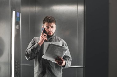 Konsantre işadamı akıllı telefonda konuşuyor ve asansörde belgeleri okuyor. Konsantre erkek ofis işçisi gazete kontrol ve telefon ile bir patron ile konuşan.