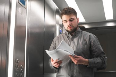 Bir sakal giyen ciddi iş adamı gri bir gömlek giyer, elinde belgeler ile asansörde duruyor ve kameraya bakarak üzerinde duruldu. Ofis asansördeki ofis işçisinin portresi