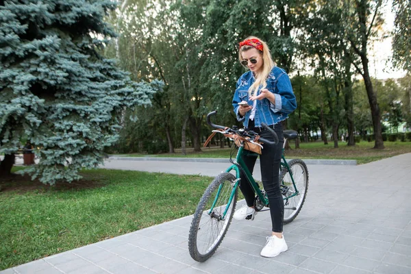 Élégante femme attrayante se tient dans un parc avec un vélo et utilise un smartphone. Fille regarde la piste cyclable sur le smartphone . — Photo