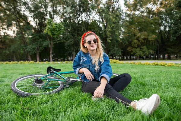 Étudiante souriante dans des vêtements élégants assis sur une pelouse verte dans un parc avec un vélo et de détente. Joyeux attrayant femme en lunettes de soleil dans un parc avec un vélo. Loisirs extérieurs — Photo