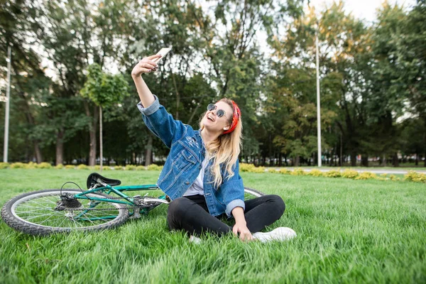 漂亮的女孩穿着时髦的衣服坐在绿色的草坪上骑自行车, 并采取自拍。年轻漂亮的女人坐在草地上, 并采取自拍与自行车在你的智能手机。在公园休息 — 图库照片