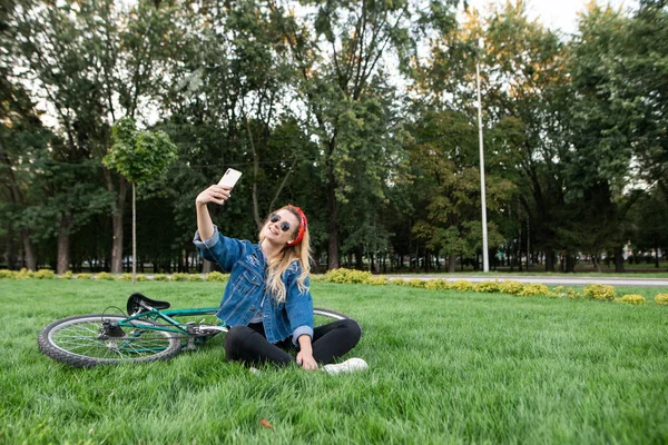 Молодая девушка сидит в траве в парке и делает селфи на смартфоне. Счастливая красивая девушка в стильной одежде и солнцезащитных очках сидит на траве в парке и делает селфи на фоне велосипеда . — стоковое фото