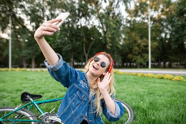Счастливая красивая девушка в стильной одежде и солнцезащитных очках сидит на траве в парке и делает селфи на фоне велосипеда. — стоковое фото