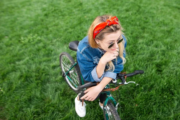 Stylowa, atrakcyjna dziewczyna na rowerze na tle zielonego trawnika. Działalność na rowerze w plenerze. Stylowa dziewczyna student z rowerem w parku — Zdjęcie stockowe