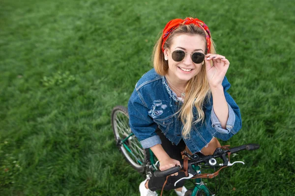 一个时尚迷人的女孩在太阳镜站在草坪上的自行车在公园里, 看着相机和微笑的肖像。骑自行车的年轻妇女在一个绿色的草地背景上的相机摆姿势. — 图库照片